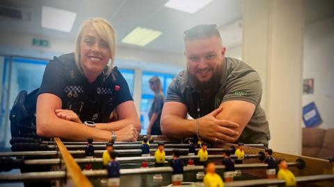 A policewoman with blonde hair leans on a football table next to a man. They both smile at the camera.