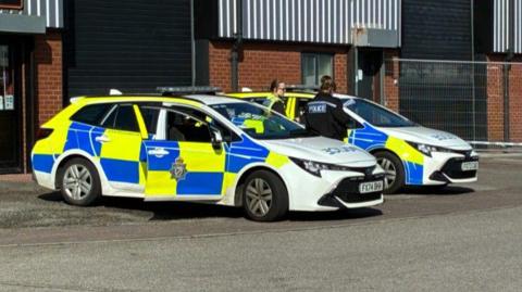 Two yellow and blue police cars are parked up in front of a building. The nearest one has its driver-side door open. Two female police officers can be seen stood in the middle of the cars.