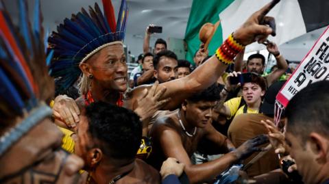 A protester, wearing a traditional headdress, raising his hand at a protest.