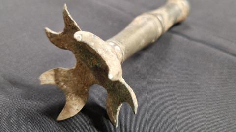The rattle is made of metal and appears heavily weathered, with visible corrosion, tarnish, and oxidation. It consists of a long cylindrical shaft or handle attached to a decorative, multi-lobed head.