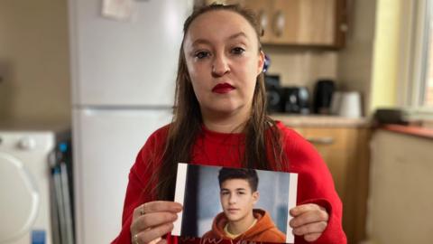 Heidi Rose Elliott, wearing a red top. She has red lipstick on and brown shoulder length hair, and is photographed in her kitchen. She is holding an AI-generated photo of her son