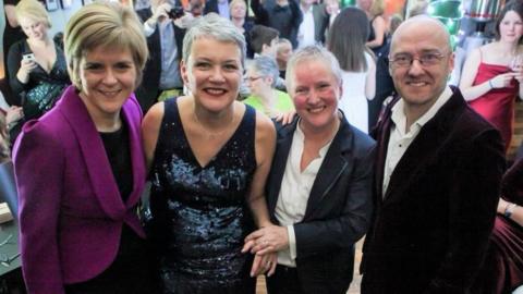 Nicola Sturgeon in purple jacket and dark blouse, Susan Douglas-Scott in a blue dress, Gerrie Douglas-Scott in a dark blue suit with white shirt and Patrick Harvie in purple velvet jacket are standing left to right at a party, there is a crowd milling about behind them. All four are smiling at camera.