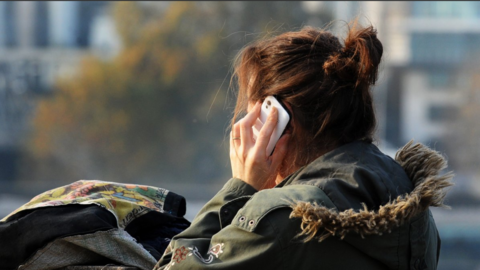 A woman with brown pulled up hair and a green coat with a hood using a mobile phone in an outdoor setting