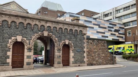 The image shows part of the Jersey General Hospital building. It is a mix of stone, brick and cladded buildings. Two yellow ambulances are parked up outside the hospital to the right.