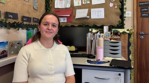 Charlotte Moore is sitting at her desk in the classroom she teaches in. She is smiling and wearing a white top. Her desk has a computer monitor and there are various items pinned up on the wall.