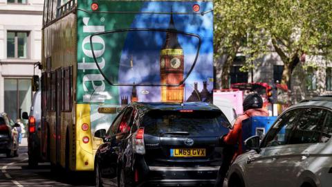 A rear view of a black Toyota Prius sitting in traffic on a city street, positioned directly behind an open-top double-decker tour bus featuring a large advertisement with an image of Big Ben and the text "Viator".