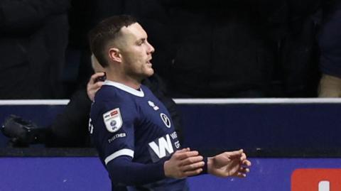 Macaulay Langstaff slides on his knees on the turf in celebration with his fists clenched, in front of the Millwall fans 