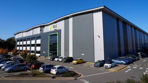 A large industrial building with light and dark grey cladding. A sign that says Hello Fresh is at the top of the building. The building is surrounded by a car park with most spaces taken up with cars.