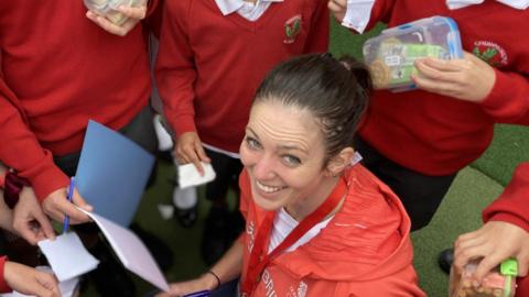 A woman in a red jacket smiles as she is surrounded by school children. 