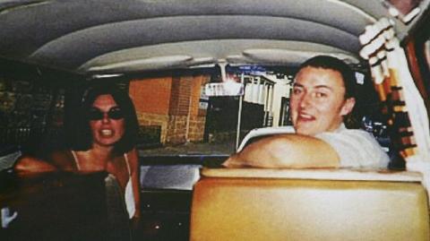 Joanne Lees and Peter Falconio sitting in a van, turning to face the person taking the photo, who is in the back seat. Lees has dark, shoulder-length hair and is wearing black sunglasses. Falconio is smiling and has short dark hair and a white t-shirt on.