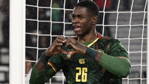 Christian Kofane, wearing a green Cameroon shirt, makes a heart shape with his hands as he celebrates a goal at the Africa Cup of Nations