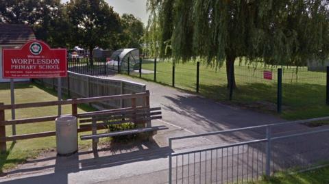 A Google maps image of the view of a primary school from a pavement outside with a red school sign and parts of the school building visible behind a large tree on the school field