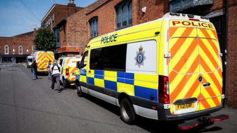 Three police vans parked on the right hand side of a road in front of a block of flats. Three people can be seen walking away from the camera. The sky is blue. 