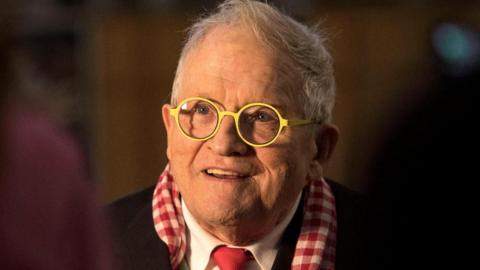 David Hockney: a man with short grey hair and round yellow glasses wearing a red check scarf and suit
