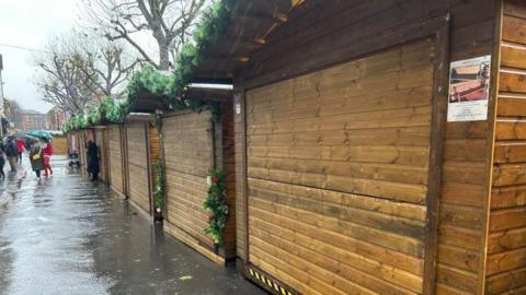 Shuttered wooden lodges with Christmas decorations around them. In the distance people in coats holding umbrellas walk away.