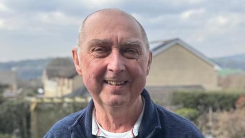 Steve Cervi, 71, who has a bald head with grey hair on the sides, is clean shaven and is wearing a blue fleece over a grey polo-neck t-shirt, smiles at the camera with a view of houses and distant hills in the background. 
