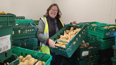 Ali Thomas is in a depot with stacks of green crates containing vegetables including potatoes and parsnips. She is wearing a hi-vis vest and has glasses. She is smiling and looking happy.