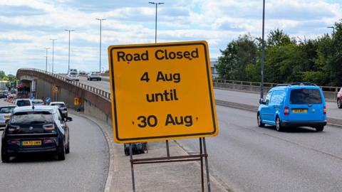 A yellow sign saying road closed 4 Aug until 30 Aug. There is a flyover in the distance with cars driving down the road.