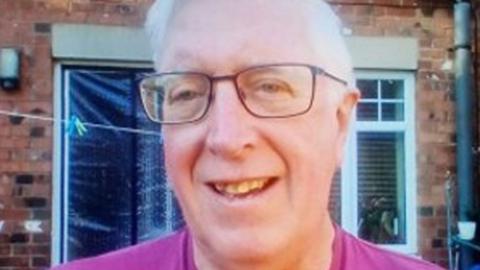 Close up of George Norwell standing outside a red brick house with white plastic windows. He is smiling and has short white hair and dark famed glasses and is wearing a pink t-shirt
