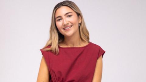 Hanan Tantush smiles for a portrait picture. She has long blonde hair and wears a red sleeveless top. She is in front of a plain white backdrop.