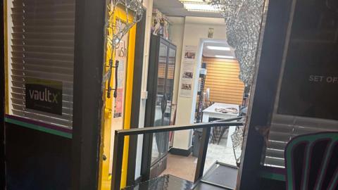 A smashed front door of a shop. Inside, there are yellow doors and what appears to be a black cabinet on the floor.