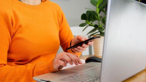 Woman in an orange jumper at a laptop computer, with a phone in her hand.