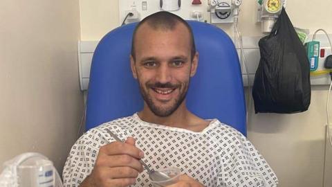 Dave Jones holding a spoon and plastic food pot and sitting on a blue hospital chair