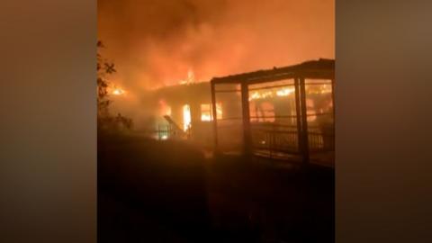 Screenshot from a video showing flames licking through the windows and doorway of a one-story building. The frame of another building sits to its right. Smoke rises high into the air and the sky is coloured orange by the flames.