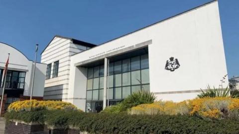 The exterior of the courts of justice building, a large white structure with large windows, you can see well-kept hedges in front.