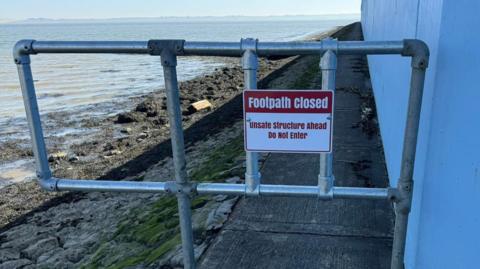 A close up of a metal barrier made from scaffolding bars with a red and white sign saying footpath closed unsafe structure ahead do not enter. It is barring entrance to a concrete path with a white wall on the right and on the left the sea wall stones drop off to the beach and the sea. The hazy sky is on the horizon. 