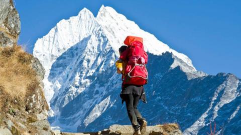 A woman carrying her toddler child in a red child carrier rucksack on her back, trekking above Namche Bazar (3,440 m), the base for trekking and mountaineering in Nepal's Solo Khumbu region