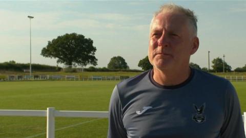 Man in grey jersey standing in front of a football pitch