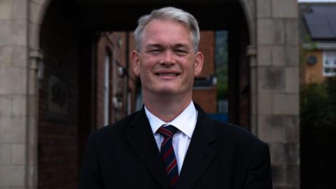 Richard Tunnicliffe, wearing a black jacket, dark blue and red tie, and white shirt, in front of a stone arch.