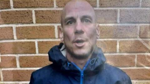 A bald man, who is wearing a dark blue coat with the hood down, has his mouth partially open as he stands in front of a wall.