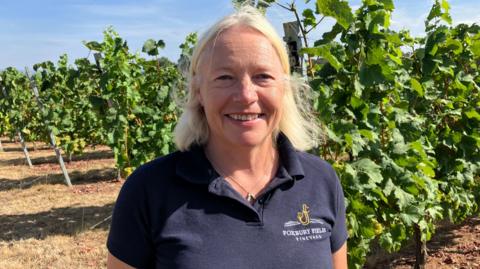 Heather Williamson from Foxbury Fields Vineyard stood in front of rows of vines of grapes. She has a bright blonde bob haircut and is wearing a navy polo shirt with the logo of the vineyard on the chest. The sky is blue and the ground is dry and brown.