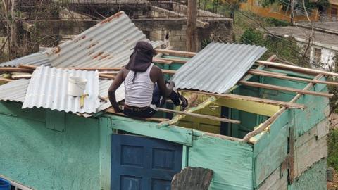 A man is sitting on the broken roof of his home trying to repair it. The house is made of wooden slabs and metal.