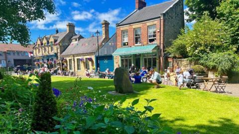 Washington on a sunny day. The sky is blue and cloudy and there are people sitting on the grass and benches at the village green outside a pub and cafe. There is bunting hanging across the green. 