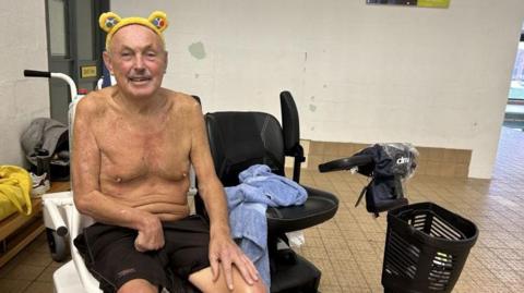 Steve Harrison sat in a hoist on poolside wearing swimming trunks and Pudsey ears while smiling into the camera. His mobility scooter is next to the hoist with a towel draped on it.