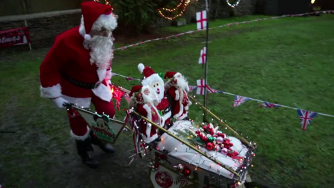 A man wearing a Father Christmas outfit drives a wheelbarrow covered in festive lights and Christmas decorations around a route in his garden, mapped out with Union Flag and England flag bunting