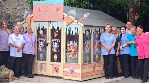 The nursing staff of Belle Vue care home are in their uniforms standing next to "Jody's Scoops of Love" which is a large shed decorated with ice cream cones. It is painted pink and cream.