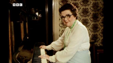 A screenshot from the BBC Look North clip that shows Peggy Fullerton playing the piano while looking into the camera. She's got short, curly brown hair and large frame glasses. She's wearing a white shirt with green beaded necklace. Behind her is a dark brown door and patterned wallpaper.