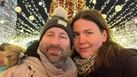 Ukrainian refugee Maria Romanenko with her face pressed close to her Mancunian partner Jez as they pose for a photograph under a large Christmas tree surrounded by Christmas lights at an outdoor street event.