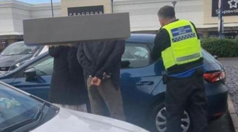 Three people next to a blue car have their backs to the camera. The signs for two stores are in the background.