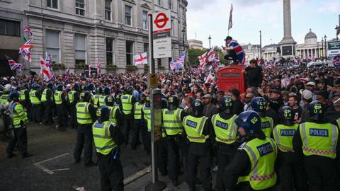 A line of police in high-visibility jackets and helmets faces a crowd waving Union Jacks and St George's Cross flags. A person draped in a flag sits atop a red telephone box near a Whitehall bus stop, with Nelson's Column in the background.
