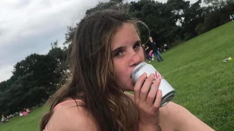 Holly Newton has long brown hair and is wearing a vest top. She is pictured sitting in a park drinking a Diet Coke.