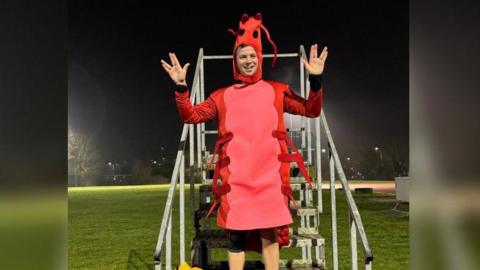 Jamie Campbell is standing in the middle of the picture, and is on some stairs. He is wearing a shrimp costume, which includes a hat with eyes on, and the body, which is pink and red, with six legs. 