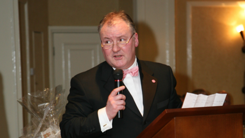 A man in a pink bow tie, with white shirt and black dinner jacket. he is wearing half moon spectacles with gold rims and appears to be speaking behind a lectern at an event. He is holding a microphone and standing in a room with yellow walls. The top of a cellophane-wrapped gift can be seen behind.