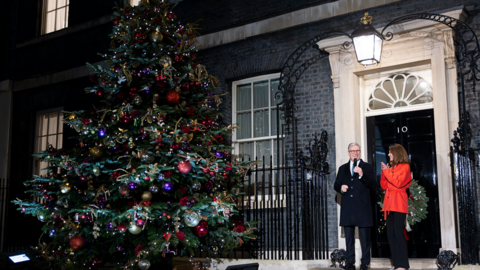 Sir Keir Starmer and a woman in a red coat stood outside the door of 10 Downing Street next to a huge Christmas tree docrated with lights, multicoloured baubles and tinsel