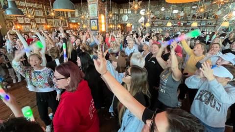 A large crowd gathered indoors at a lively event, with people packed closely together and raising colourful light sticks, surrounded by warm lighting, framed pictures on the walls and hanging lamps in a busy bar‑style venue.