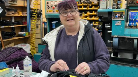 Jackie Ashcroft, who has dyed purple hair cut into a fringe, glasses and is wearing a grey gilet over a purple jumper, smiles at the camera from behind a turquoise shop counter.